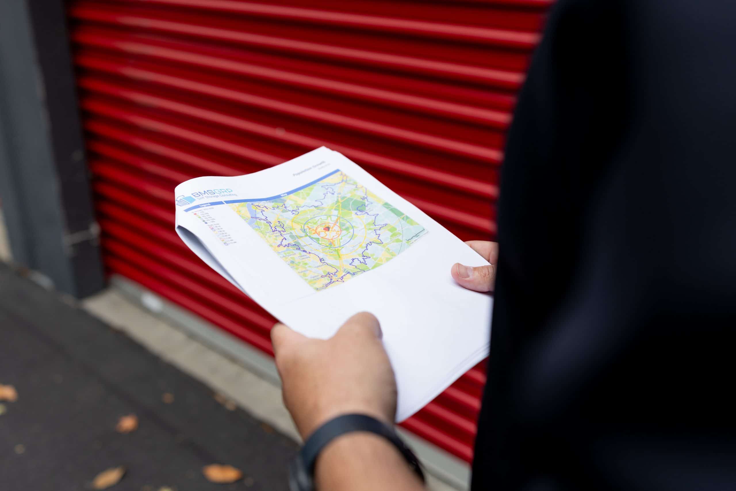 BMSGRP professional reviewing a self-storage market analysis map in front of a storage facility with red roll-up doors.
